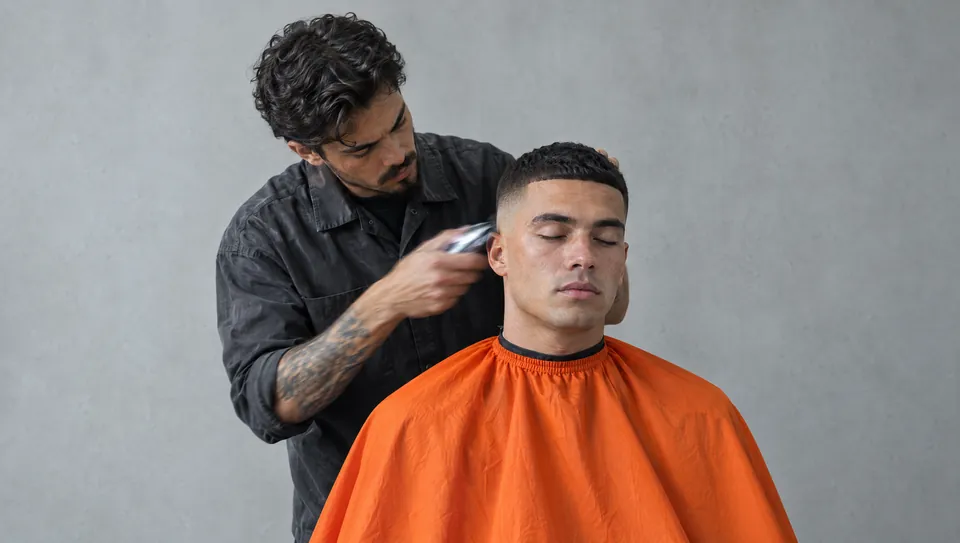 A barber in a charcoal shirt details a skin fade on a client in an orange cape, eyes closed, grey studio backdrop
